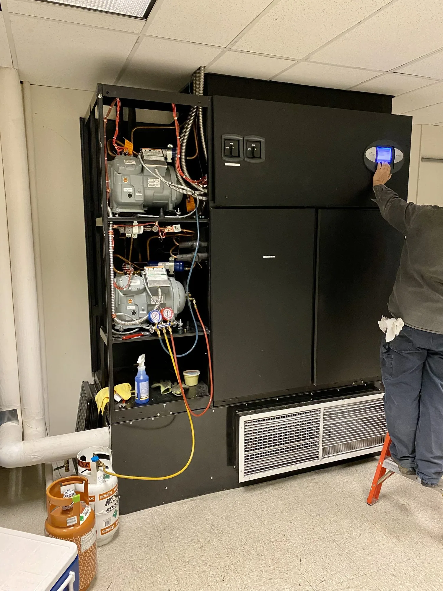 A JCD technician at an opened cooling unit in a commercial mechanical room, gauges connected